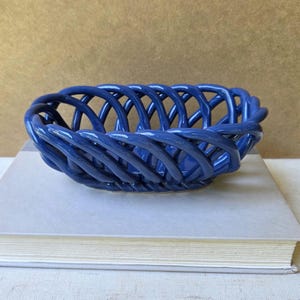 Puede incluir: Una cesta de cerámica azul profundo brillante con un intrincado diseño de celosía de tejido abierto. El cuenco de forma ovalada está construido con hebras de cerámica redondeadas e interconectadas que forman una estructura continua y lisa.