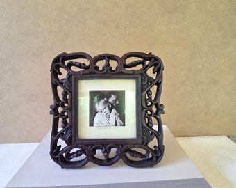 May include: Ornate dark brown picture frame with a square photo of a parent and child. The frame has a detailed, scrolling design. The photo is centered within the frame, which is resting on a striped surface.