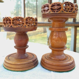 May include: Two wooden candle holders with intricate carved designs. The candle holders are made of a light brown wood and have a round base, a central stem, and a decorative top with openwork patterns.