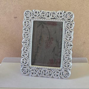 May include: A rectangular silver-toned picture frame featuring an intricate, raised relief pattern of scrolling vines and leaves along the border. The frame surrounds a dark grey fabric insert decorated with a subtle, muted red floral print design.