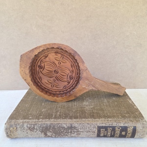 May include: A vintage wooden butter mold with a handle, featuring a detailed floral and geometric design carved into the circular mold. The mold is a light brown color and rests on a worn, antique book.