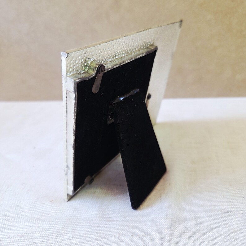 May include: A small, silver-colored picture frame with a textured border. The frame has a black velvet backing and a hinged easel stand. The frame is designed to stand upright on a flat surface.