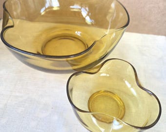 May include: Two amber-colored glass bowls with a unique, wavy rim design. The bowls are nested, with one larger than the other. The glass has a smooth, transparent appearance, and the bowls are likely for serving or display.