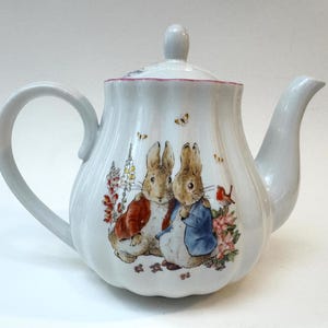 May include: White ceramic teapot with a lid and handle. The teapot features a colorful illustration of two rabbits in clothing, surrounded by flowers and butterflies. The spout and handle are curved.