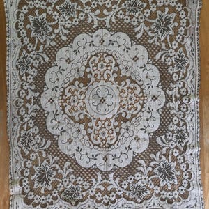 May include: A square white lace tablecloth with a brown floral pattern. The lace has a scalloped edge.