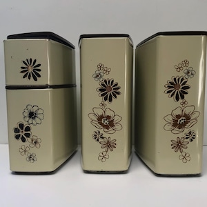 May include: A set of three vintage beige metal canisters with dark brown lids. Each canister has a floral design in brown and black. The canisters are rectangular with rounded corners, ideal for kitchen storage.