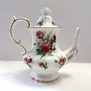 May include: White porcelain teapot with a gold trim and a floral design featuring pink roses. The teapot has a lid with a knob in the shape of a rose.