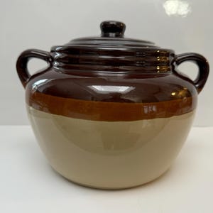 May include: A brown and beige ceramic bean pot with two handles and a lid. The pot has a glazed finish and is decorated with a brown band around the middle.