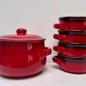 May include: A red ceramic pot with a lid and two handles, beside a stack of four small red bowls. The bowls and pot have a glossy finish and a dark brown rim.