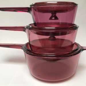 May include: Set of three purple glass cookware with lids. The cookware is nested together with the handles extending out.