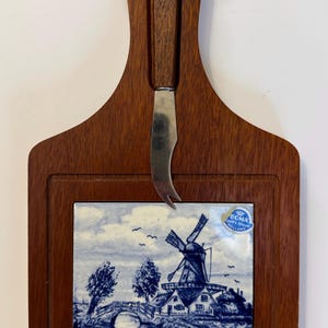 Peut inclure: Une planche à fromage en bois avec une tuile de Delft bleue et blanche représentant un moulin à vent et un pont. La planche est dotée d'un couteau intégré et d'une boucle de suspension en cuir. La tuile est marquée "Boma Delft's Blue Holland".