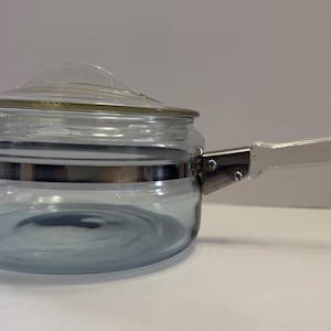 May include: A clear glass saucepan with a silver metal band around the middle and a clear glass handle. The saucepan has a lid.