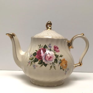 Peut inclure: Une théière blanche avec un motif floral composé de roses roses, jaunes et blanches. La théière est ornée d'une bordure dorée et d'un couvercle.