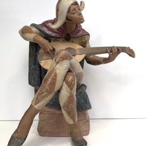 May include: A porcelain figurine of a jester playing a lute, wearing a colourful jester costume with a red and white hat. The figurine is sitting on a stone base.