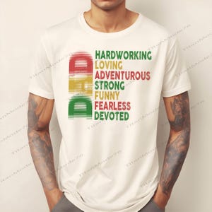 May include: A white t-shirt with the word "DAD" in red, yellow, and green, with the words "HARDWORKING, LOVING, ADVENTUROUS, STRONG, FUNNY, FEARLESS, DEVOTED" in green and red. The shirt is made of a soft material.