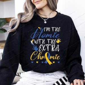 May include: Black sweatshirt with the text "I'M THE Homie WITH THE EXTRA Chromie" in white and blue script. A yellow and blue ribbon graphic is below the text, with blue hearts.