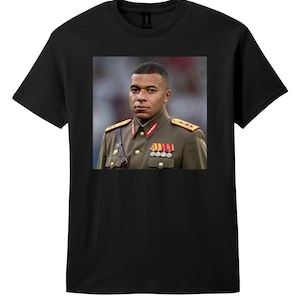 May include: A black short-sleeved t-shirt featuring a centered graphic print of a person with short dark hair wearing an olive green military-style uniform with gold buttons, red collar tabs, and multiple silver medals pinned to the chest.