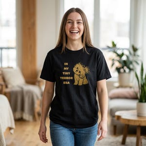 以下が含まれることがあります： 「IN MY TINY TERROR ERA」の文字と犬のイラストが描かれた黒いTシャツ。犬は薄茶色でふわふわしています。Tシャツは、青いジーンズを履いた人が着ています。