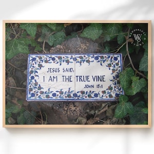 May include: Framed artwork featuring a rectangular tile with the text "JESUS SAID: I AM THE TRUE VINE JOHN 15:1". The tile has a blue and white floral border. The artwork is set against a backdrop of green foliage.