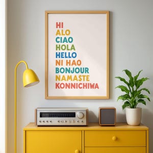 May include: A framed wall art print featuring the words HI, ALO, CIAO, HOLA, HELLO, NI HAO, BONJOUR, NAMASTE, and KONNICHIWA in various colors, arranged in a vertical list format on a white background within a light wood frame.
