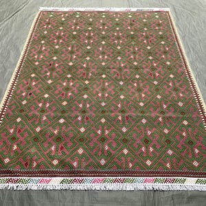 May include: A rectangular rug with a geometric pattern in shades of green and pink. The rug has a cream-colored border with a fringe along the bottom edge. The pattern consists of repeating diamond and square shapes.