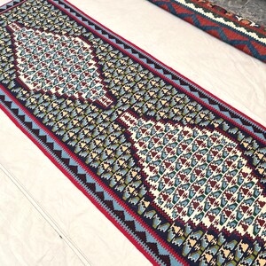 May include: A long rectangular runner rug featuring a repeating geometric pattern in shades of dark blue, cream, light blue, and deep red. The design includes two prominent central diamond-shaped motifs and a decorative border with a zigzag edge.