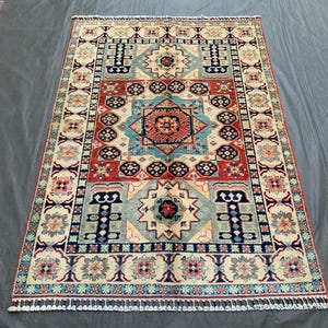 May include: Rectangular area rug with a detailed geometric pattern. The rug features a central design in red, blue, and cream, surrounded by borders with repeating motifs. The rug has a fringe along the edges.
