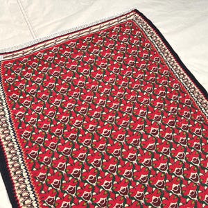 May include: A rectangular woven rug featuring a repeating floral pattern in shades of red, dark green, and cream. The rug is bordered by a dark black edge and a decorative cream and black geometric trim, finished with short white fringe at the ends.