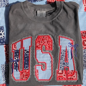 May include: A charcoal gray t-shirt with the word "USA" embroidered in red, white, and blue patchwork. The letters are outlined with a white lace-like trim and accented with blue bows. The shirt is folded on a patchwork quilt.