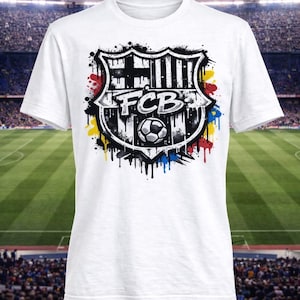 May include: White t-shirt featuring the FC Barcelona logo. The logo is a shield design with the letters "FCB" and a soccer ball, with colorful paint splatter accents. The shirt is displayed in front of a stadium background.