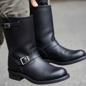 Puede incluir: Un par de botas de moto de cuero negro. Las botas tienen una altura media de pantorrilla, una puntera redondeada y una suela resistente. Cada bota presenta una correa con hebilla alrededor del tobillo y otra cerca de la parte superior. Las botas son sostenidas por una persona.