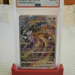 Puede incluir: Una carta Pok&eacute;mon Mewtwo VSTAR calificada del set 2023 Pok&eacute;mon SWSH Crown Zenith. La carta est&aacute; encapsulada en un soporte protector transparente y se muestra en un soporte rojo. La carta tiene una colorida ilustraci&oacute;n de Mewtwo.