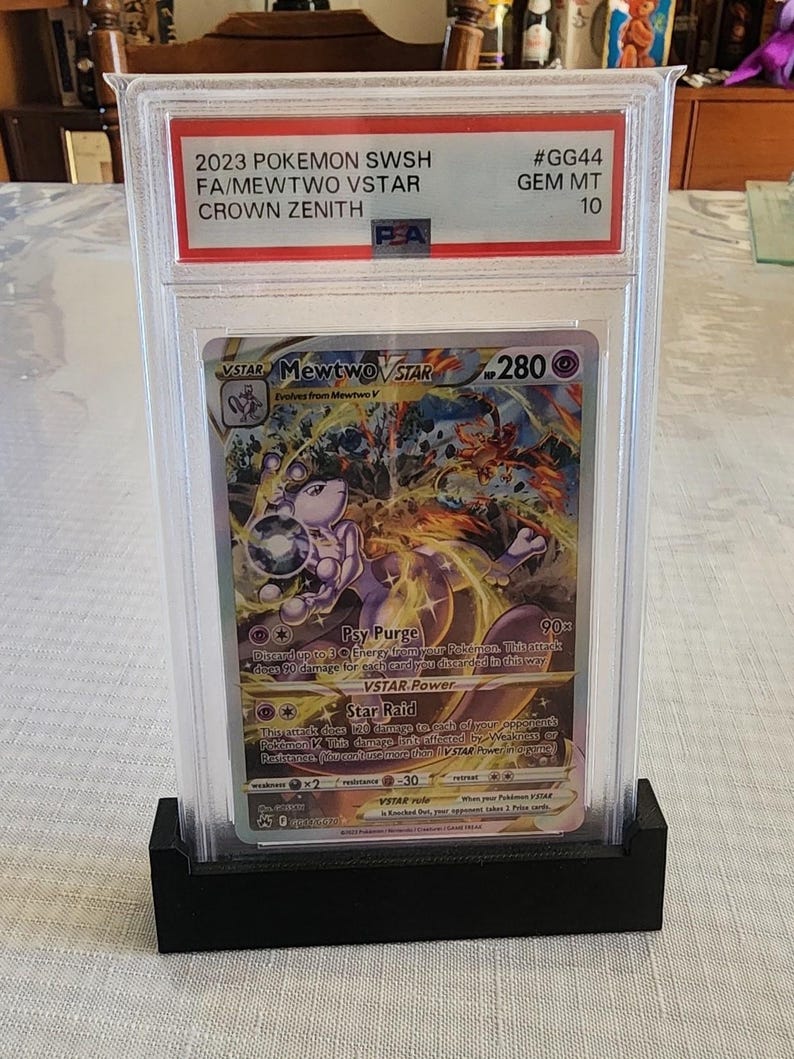 Puede incluir: Una tarjeta Pok&eacute;mon de 2023, calificada, con Mewtwo VSTAR del conjunto Crown Zenith. La tarjeta est&aacute; encerrada en un soporte protector con una base negra. La tarjeta contiene el texto "2023 POKEMON SWSH FA/MEWTWO VSTAR CROWN ZENITH".