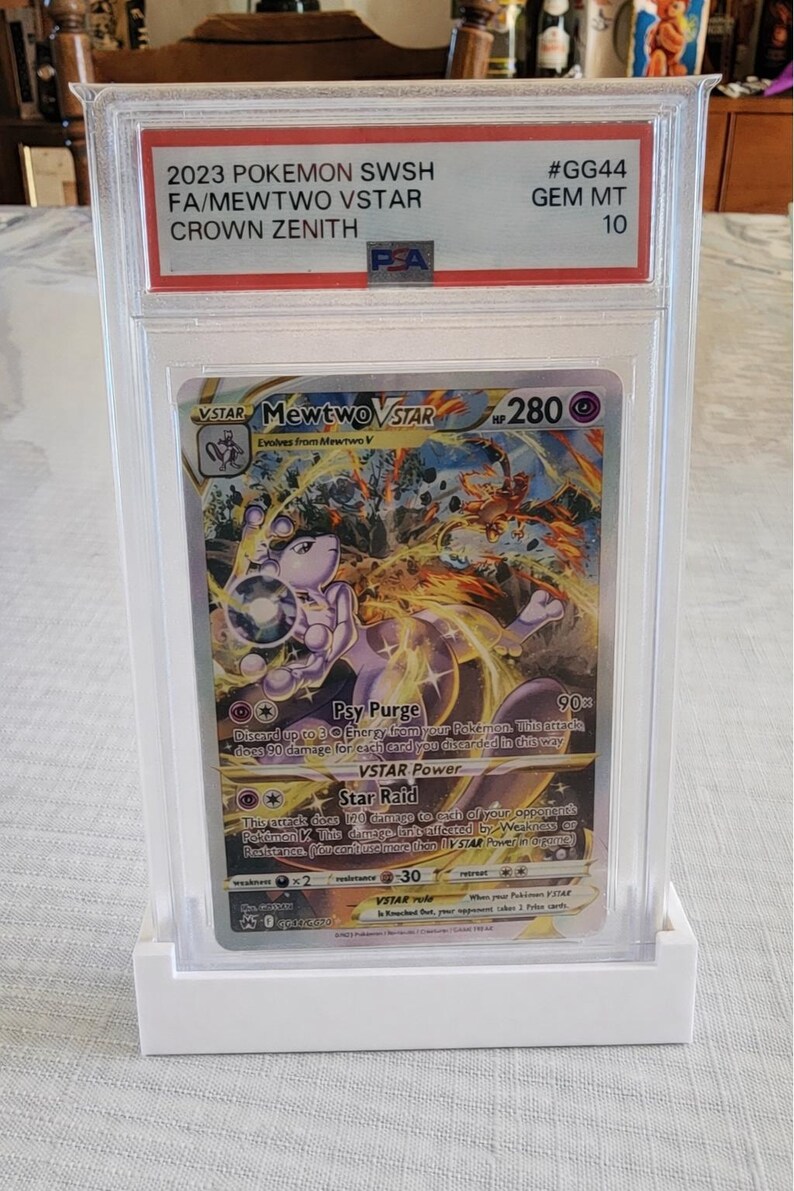 Puede incluir: Una carta de Pok&eacute;mon calificada con Mewtwo VSTAR del conjunto Crown Zenith. La carta est&aacute; encerrada en un soporte protector transparente con una base blanca. El texto de la tarjeta incluye "Psy Purge" y "Star Raid". La calificaci&oacute;n es GEM MT 10.