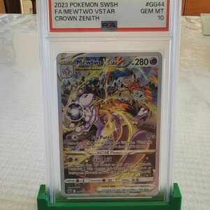 Puede incluir: Una carta Pok&eacute;mon de Mewtwo VSTAR del set Crown Zenith, calificada y protegida en un estuche de pl&aacute;stico transparente. La carta se muestra en un soporte verde. El texto de la carta incluye "2023 POKEMON SWSH FA/MEWTWO VSTAR CROWN ZENITH".