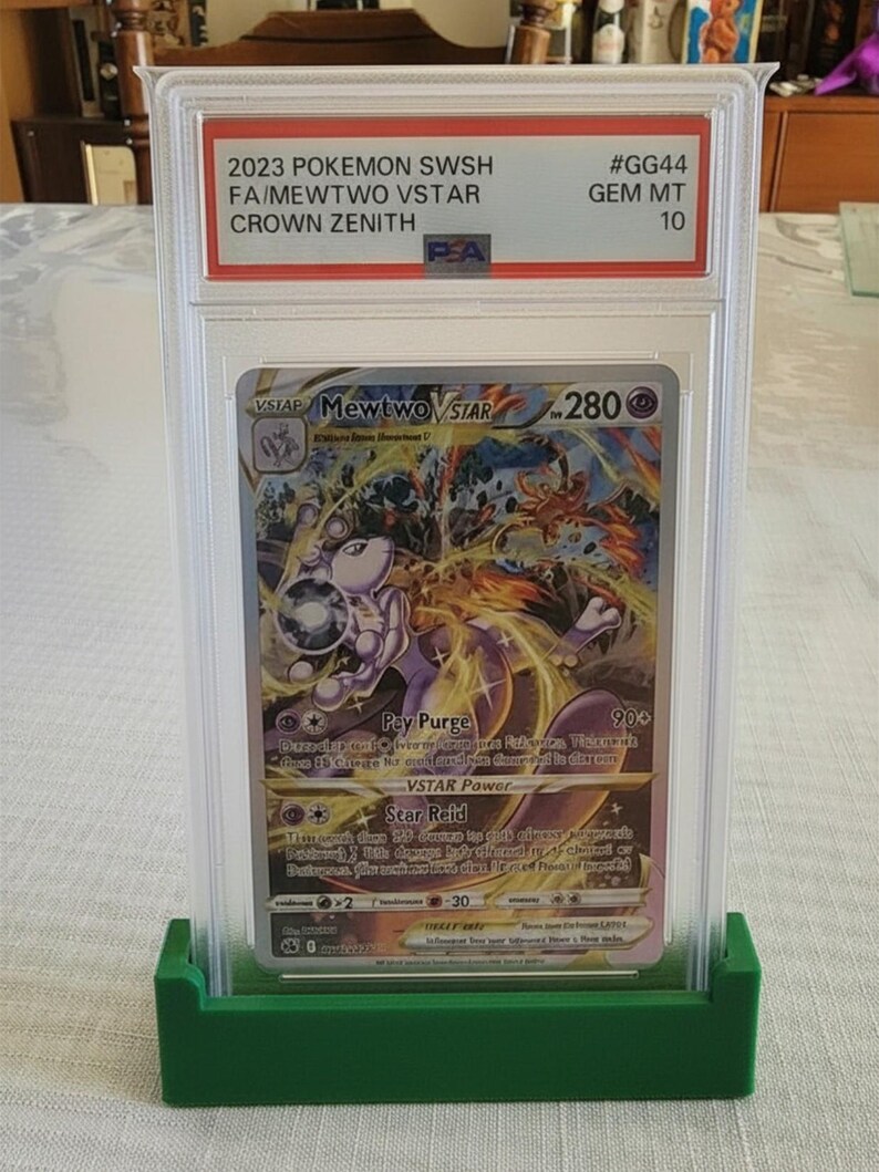 Puede incluir: Una carta Pok&eacute;mon de Mewtwo VSTAR del set Crown Zenith, calificada y protegida en un estuche de pl&aacute;stico transparente. La carta se muestra en un soporte verde. El texto de la carta incluye "2023 POKEMON SWSH FA/MEWTWO VSTAR CROWN ZENITH".