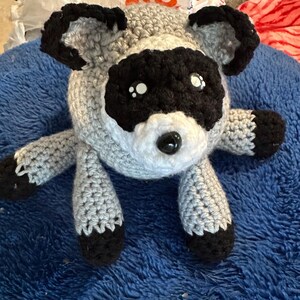May include: A handmade crochet raccoon plush toy featuring a round grey body, a white muzzle, and black patches around the eyes. The stuffed animal has black ears, black paws, and a small black plastic nose with two glossy black and white eyes.