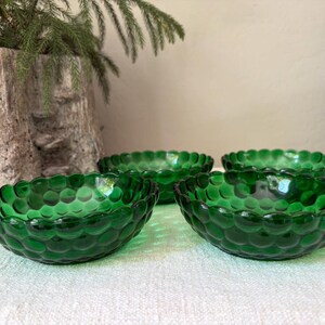 May include: A set of four small, round, emerald green glass bowls featuring a raised hobnail or bubble texture on the exterior surface. Each bowl has a scalloped rim and a translucent finish, arranged in a cluster on a textured white surface.
