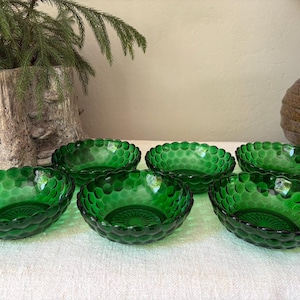 May include: A set of six small, round, emerald green glass bowls. Each bowl features a raised, circular hobnail pattern covering the exterior surface and a sunburst or starburst design embossed on the interior base of the glass.