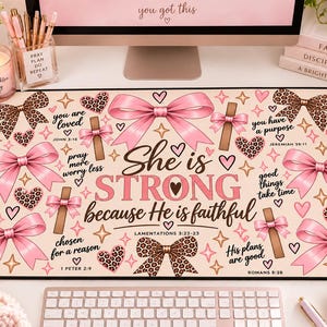 Puede incluir: Una alfombrilla de escritorio rectangular con fondo rosa claro, decorada con un patrón de lazos rosas, corazones con estampado de leopardo y pequeñas cruces. El texto dice: She is STRONG because He is faithful, junto a frases inspiradoras.