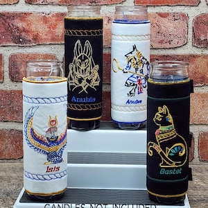 May include: Four glass candle holders with embroidered fabric sleeves featuring Egyptian deities: Anubis, Isis, and Bastet. The sleeves are black and white with gold and blue accents. The text "Anubis", "Isis", and "Bastet" are visible on the sleeves. The bottom of the image reads "CANDLES NOT INCLUDED".