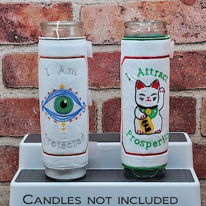 May include: Two glass candle holders with white fabric sleeves. One sleeve features an embroidered eye design and the words "I Am Protected." The other sleeve has a lucky cat design and the words "I Attract Prosperity."