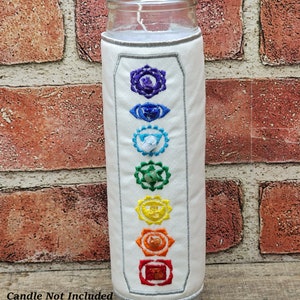 May include: A clear glass candle with a white candle inside. The candle is wrapped in a white fabric sleeve embroidered with colourful chakra symbols. The text "Candle Not Included" is at the bottom.