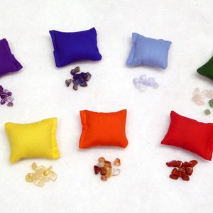 May include: Seven small, square pillows in a rainbow of colours: purple, blue, light blue, green, yellow, orange, and red. Each pillow is paired with a small pile of gemstones in complementary colours. The arrangement is on a white surface.