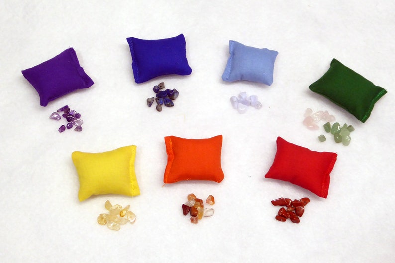 May include: Seven small, square pillows in a rainbow of colours: purple, blue, light blue, green, yellow, orange, and red. Each pillow is paired with a small pile of gemstones in complementary colours. The arrangement is on a white surface.