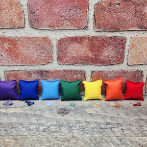 May include: A rainbow of small, square, fabric pillows in purple, blue, cyan, green, yellow, orange, and red. The pillows are arranged in a row, with various gemstones and a coin scattered in front of them. The background is a brick wall.