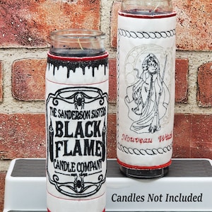 May include: Two glass candle holders with decorative fabric sleeves. One sleeve features a black and white label with the text "The Sanderson Sisters Black Flame Candle Company". The other sleeve has a witch design and the text "Nouveau Witch". Both have red trim.