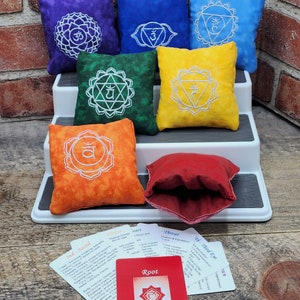 May include: Six square fabric pouches in purple, dark blue, light blue, green, yellow, and orange, each featuring white embroidered chakra symbols. A red card labelled Root Chakra and several white informational cards are placed in the foreground.