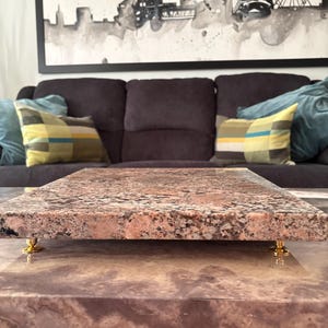 May include: A rectangular, pink and brown granite serving tray with gold-coloured feet. The tray is displayed on a matching granite surface. The background includes a dark grey sofa with decorative cushions and a black and white cityscape artwork.