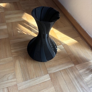 Könnte beinhalten: Eine schwarze, dekorative Vase mit einem einzigartigen, gerippten Design. Die Vase hat einen breiten, abgerundeten Boden, der sich zur Mitte hin verjüngt, bevor er sich oben wieder ausdehnt. Sie besteht aus einem festen Material und steht auf einem Holzboden.